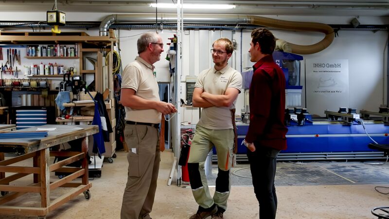 Drei Männer stehen in einer Werkstatt mit Maschinen und Werkzeugen, zwei tragen helle Poloshirts, einer ein rotes Hemd.