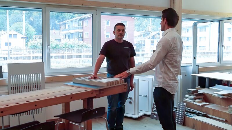 Zwei Männer stehen in einer Werkstatt mit großen Fenstern, einer trägt ein weißes Hemd, der andere ein schwarzes T-Shirt, sie unterhalten sich neben einem Holztisch mit Werkstücken