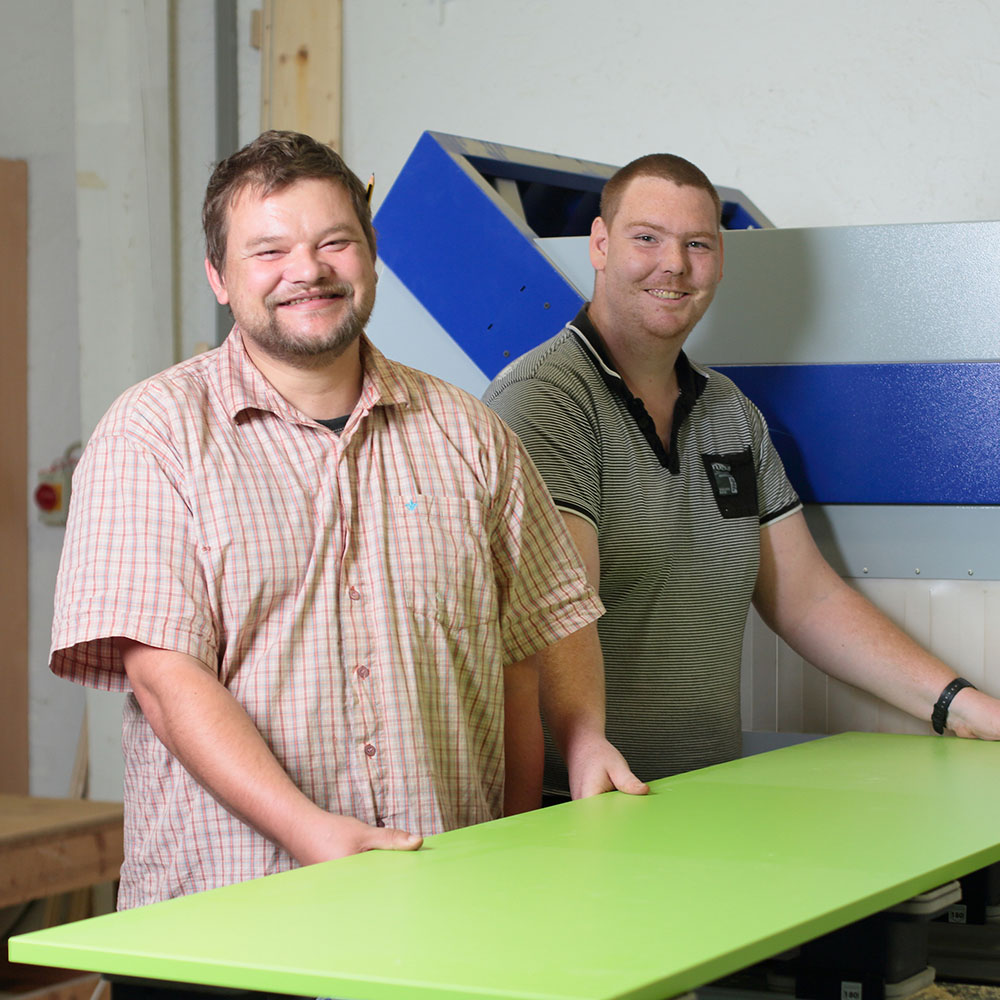 Zwei Männer stehen an einer grünen Tischplatte in einer Werkstatt, eine blaue Maschine im Hintergrund