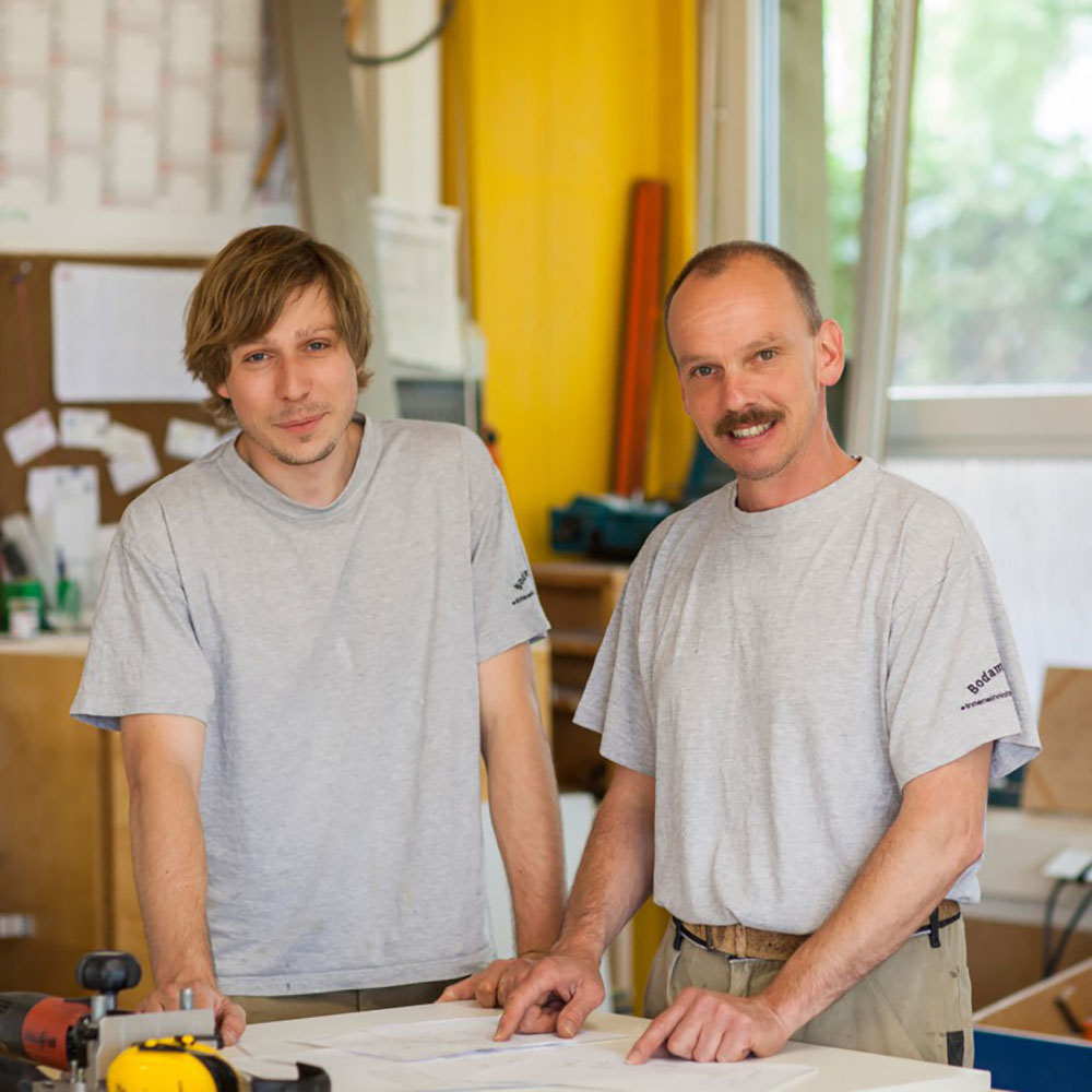 Zwei Männer in grauen T-Shirts stehen an einem Tisch mit Bauplänen in einem hellen Raum mit gelber Wand und Fenster