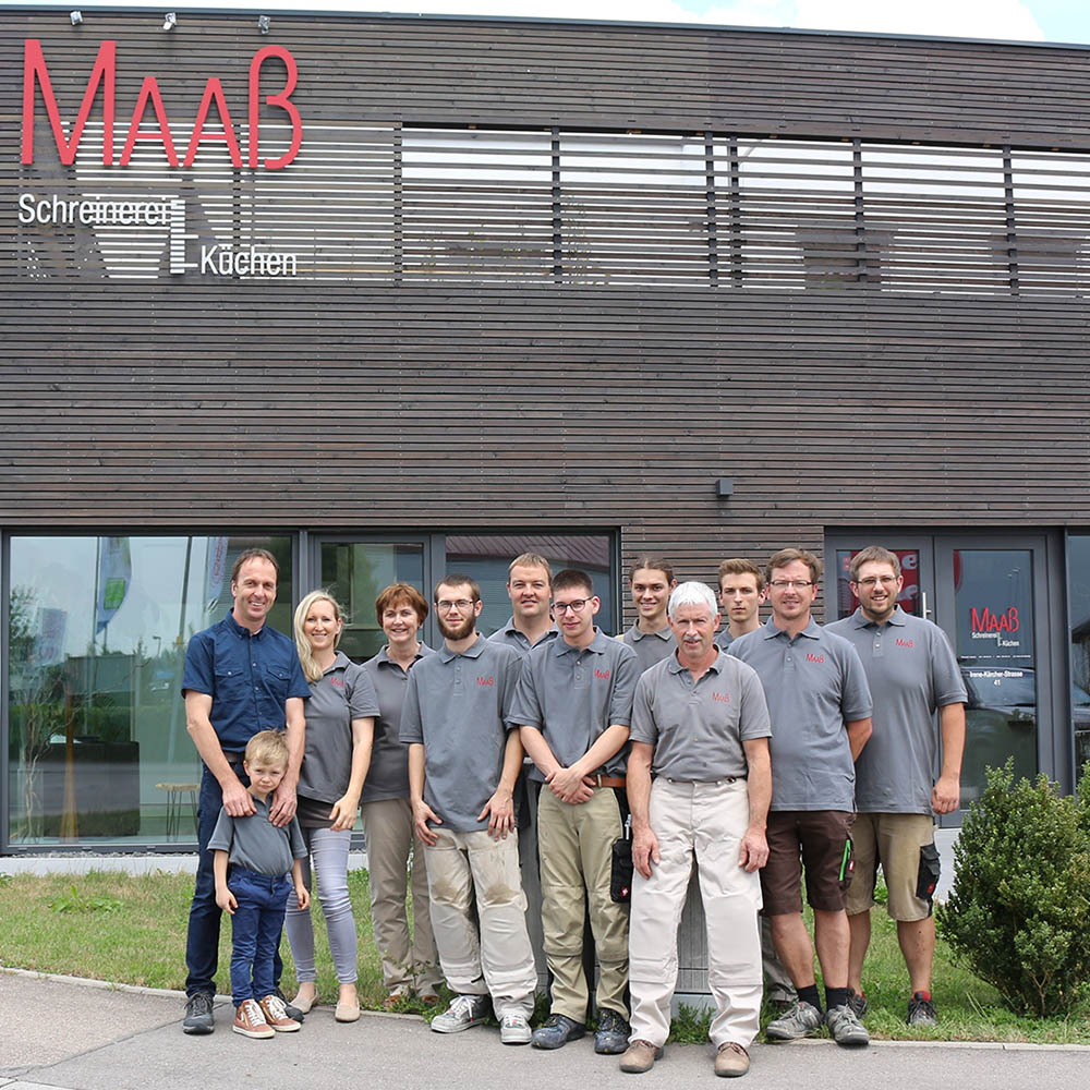 Team der Schreinerei Maaß steht vor ihrem Firmengebäude mit Holzfassade und rotem Logo