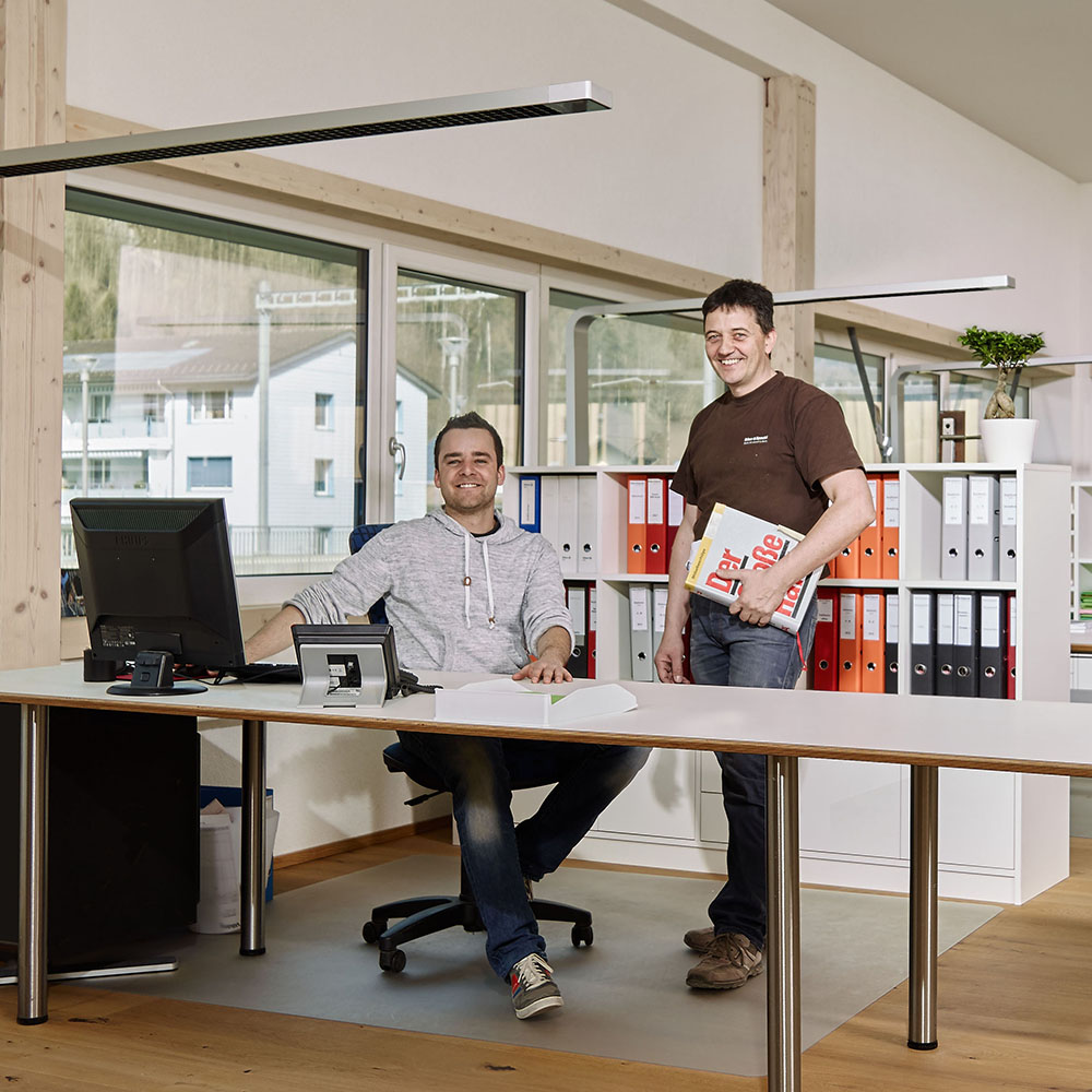 Zwei Männer im Büro, einer sitzt am Schreibtisch mit Computer, der andere steht mit Ordner in der Hand vor einem Regal mit bunten Aktenordnern