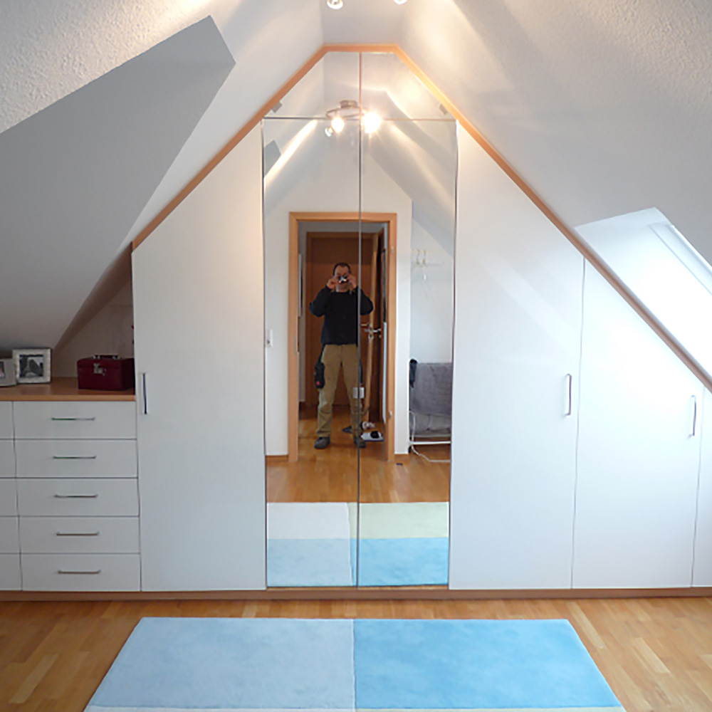 Einbauschrank unter Dachschräge mit weißen Türen, mittig zwei Spiegeltüren, links Kommode mit Schubladen, heller Holzboden, blauer Teppich