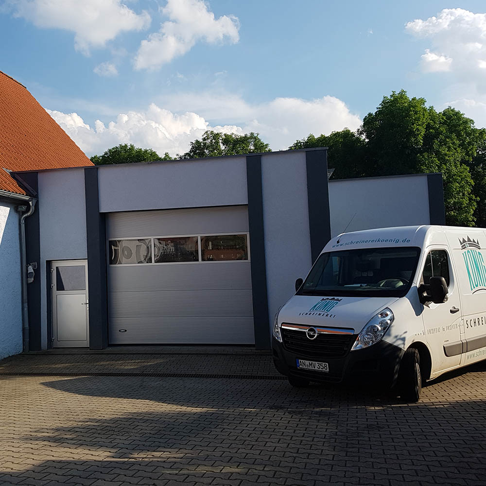 Weißer Opel Transporter mit Aufschrift 'Schreinerei Koenig' vor grauer Werkstatt mit Rolltor und Tür, blauer Himmel mit Wolken