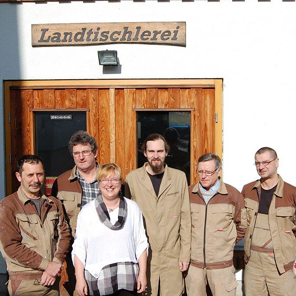 Sechs Personen stehen vor einem Holztür-Eingang mit dem Schild 'Landtischlerei' darüber, fünf tragen beige Arbeitskleidung, eine Frau trägt weißen Pullover und kariertes Hemd.