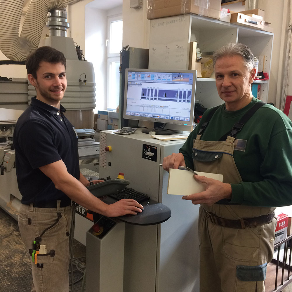 Zwei Männer in Werkstatt mit Maschinenkleidung bedienen Computer an einer CNC-Maschine mit technischer Software auf Bildschirm