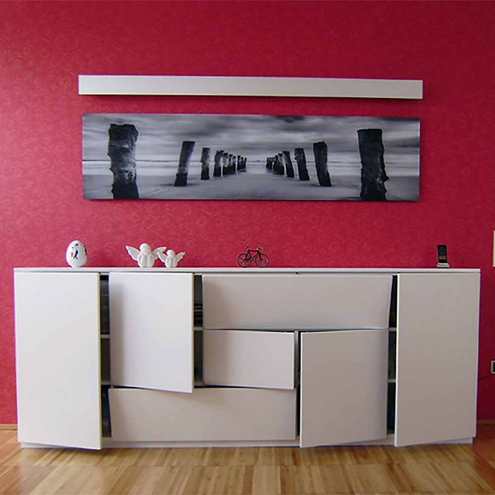 Weißes Sideboard mit offenen Türen vor roter Wand, darüber schwarz-weißes Panoramafoto von Pfählen im Wasser